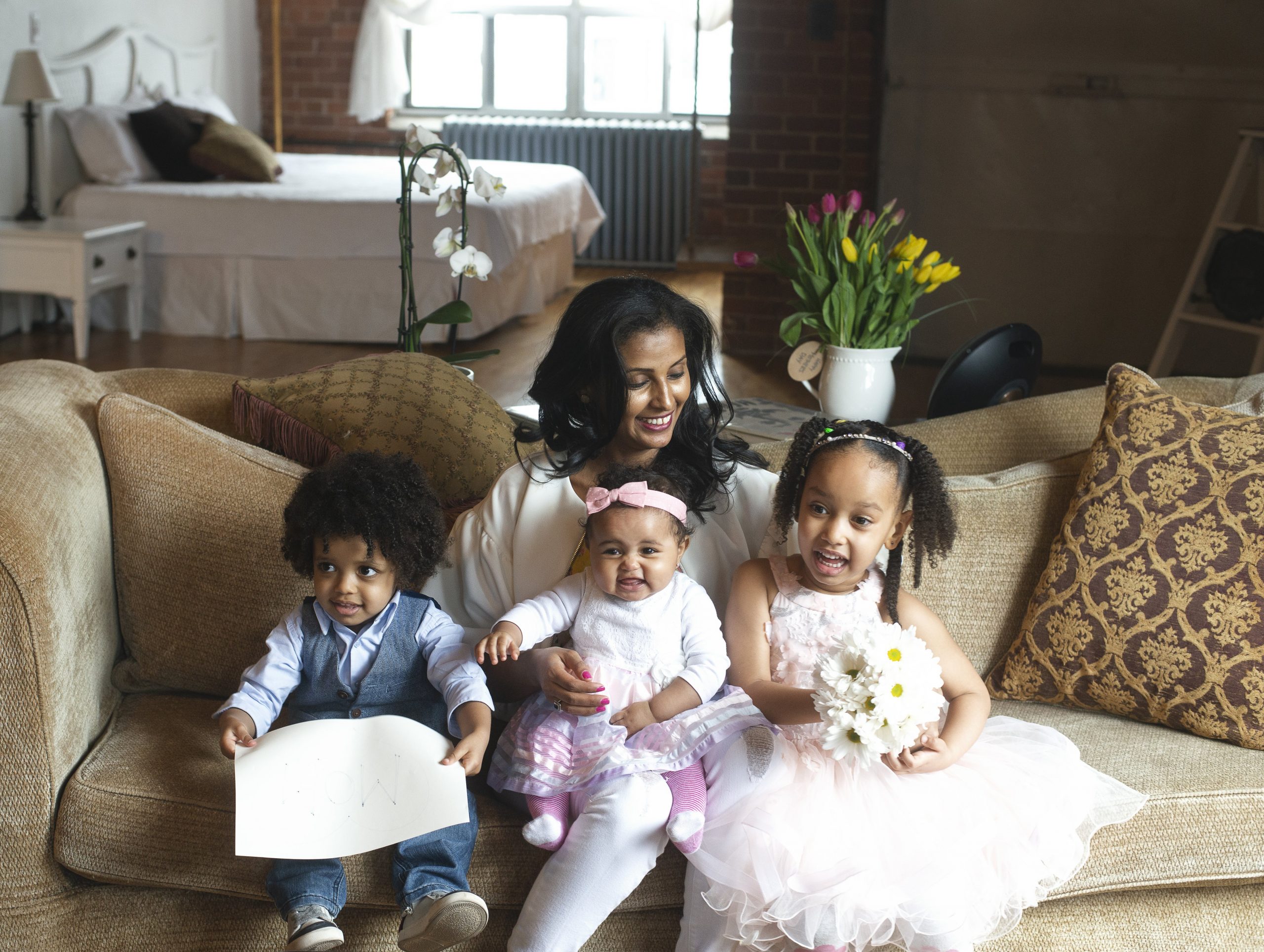 family-dressed-up Mother with three young children, a young toddler in a suit on the left, a baby with a pink bow in the middle of the mother's lap, and a young girl in a pink dress holding flowers on the right.